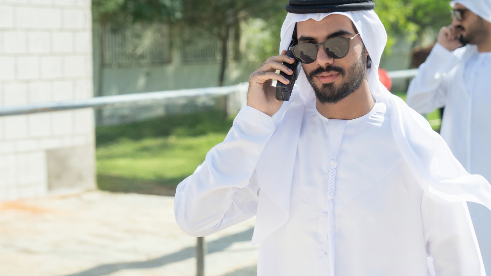 An Emirati businessman in a white kandura and ghutra taking a call on his mobile phone outdoors in the UAE, with a second Emirati colleague on a call behind him