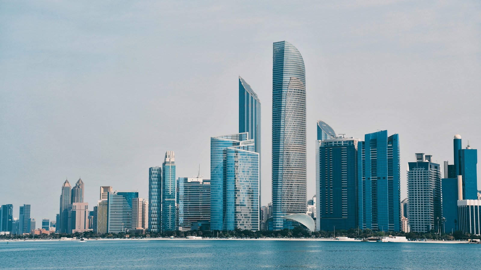 The Abu Dhabi Corniche skyline seen across calm water on a hazy day, with the curved ADNOC headquarters tower rising above a dense row of glass office and residential high-rises