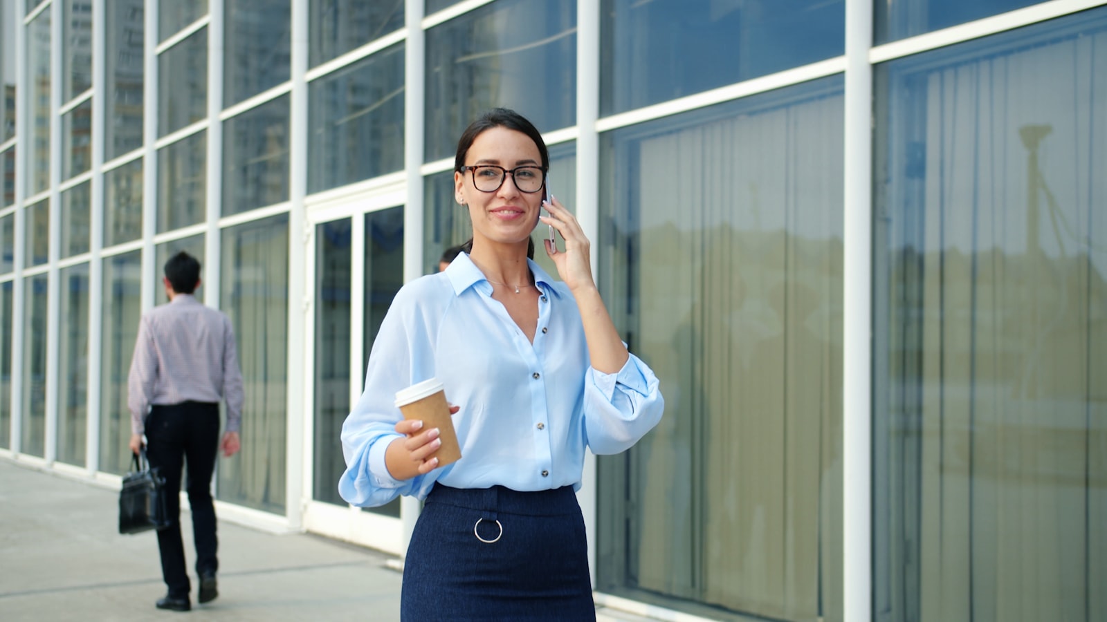 A businesswoman in a light blue shirt taking a call on her mobile phone with a takeaway coffee in her hand as she walks alone past the glass facade of a modern office building