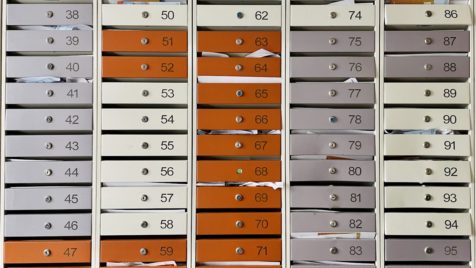 A wall of numbered apartment letterboxes in cream, grey and orange, with envelopes and folded post poking out from behind several of the flaps