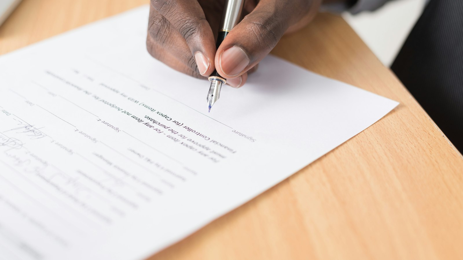 Close-up of a hand signing a printed contract with a silver fountain pen, the nib touching the signature line above two signatures already completed on the form