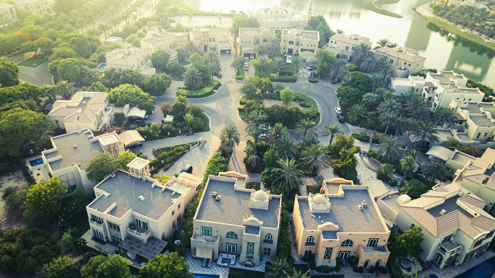 Aerial view of a Dubai villa community at golden hour, showing rows of repeated cream-coloured villa rooftops arranged around palm-lined crescent streets beside a lake