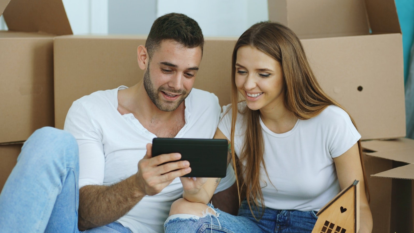 Smiling young couple sitting among moving boxes browsing property listings together on a tablet, with a small wooden house cutout in hand