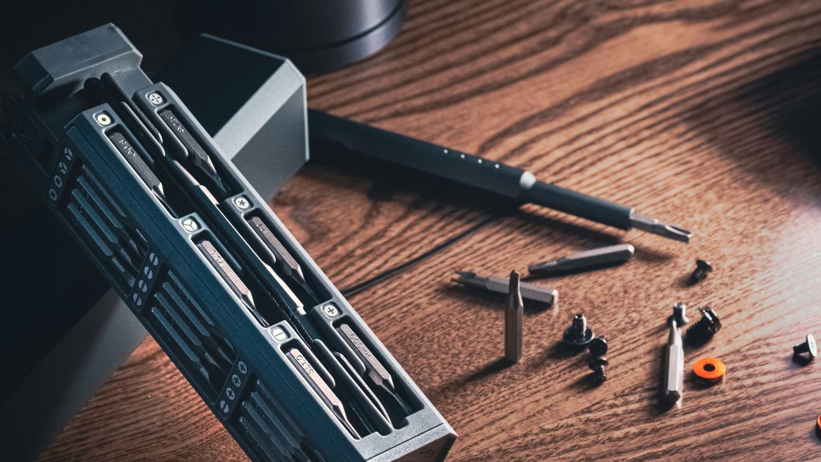 A compact precision screwdriver kit in a slim grey magnetic case, its labelled bits slotted in neat rows, beside a slim screwdriver and loose bits and screws on a warm walnut desk