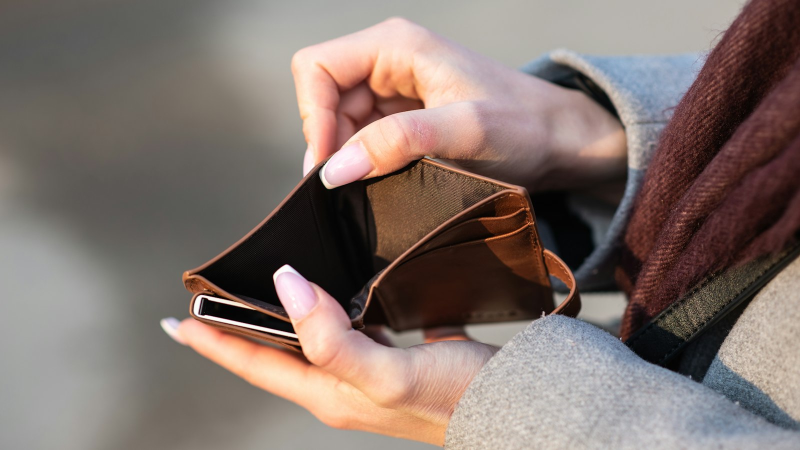 Close-up of a person's hands holding open a completely empty brown leather wallet, shot in bright daylight against a soft grey background
