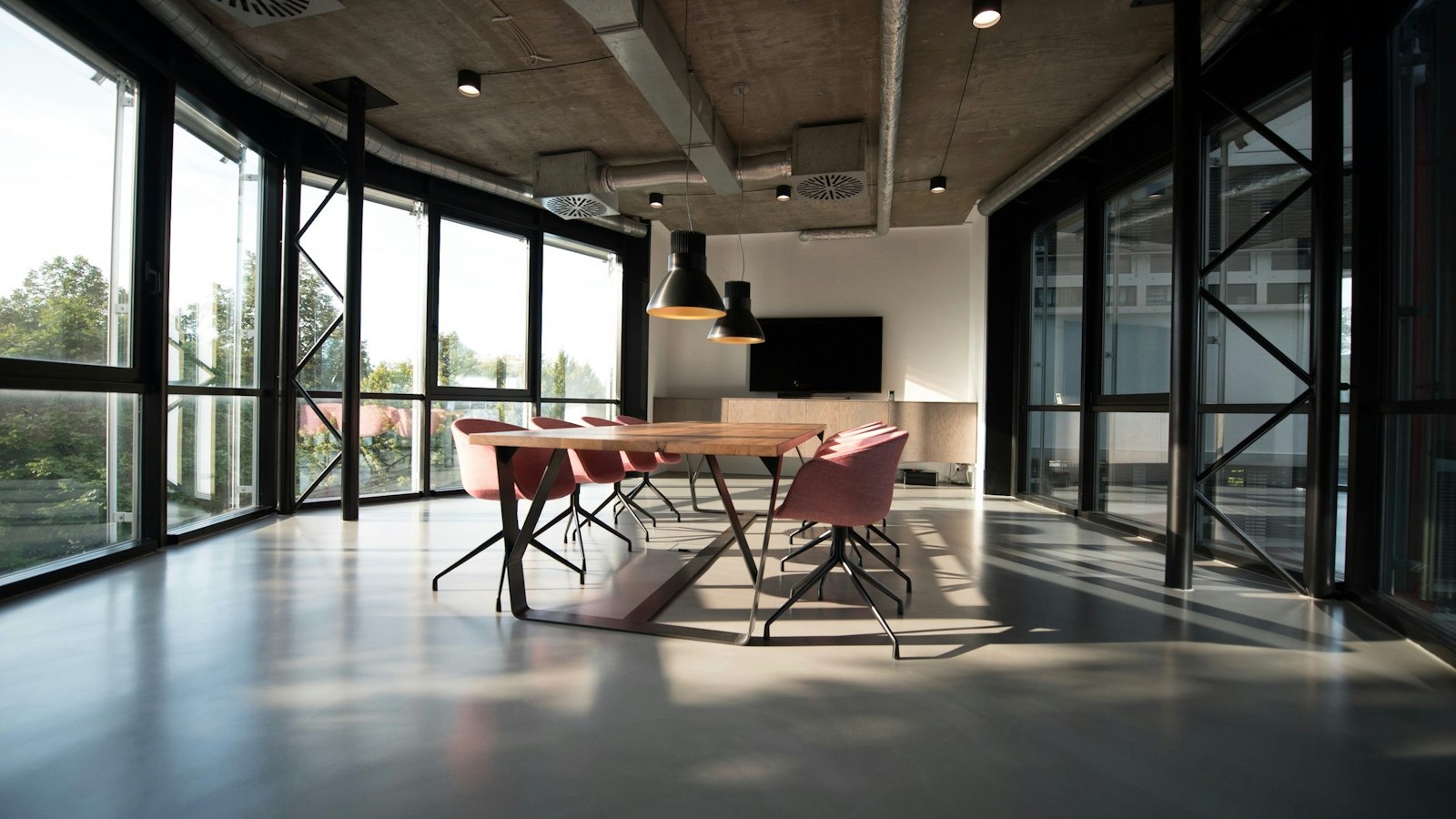 An empty commercial office floor with bare concrete soffit and exposed ducting, floor-to-ceiling black-framed glazing and a polished screed floor, holding a single timber meeting table surrounded by six vacant chairs