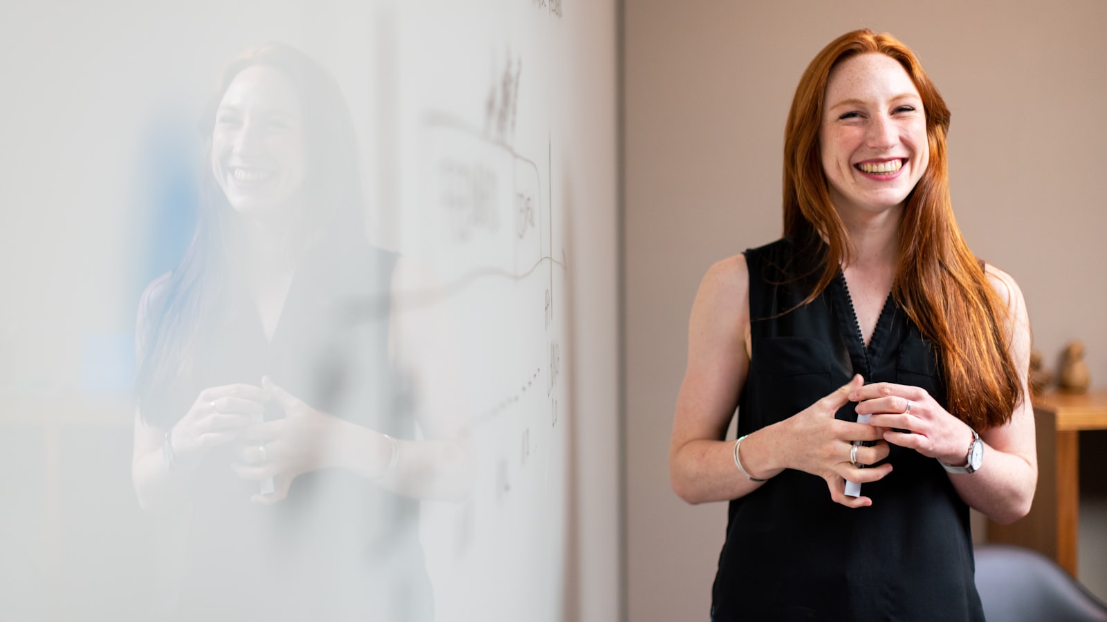 A smiling young real estate agent holding a marker at an office whiteboard, planning her first client pipeline during onboarding