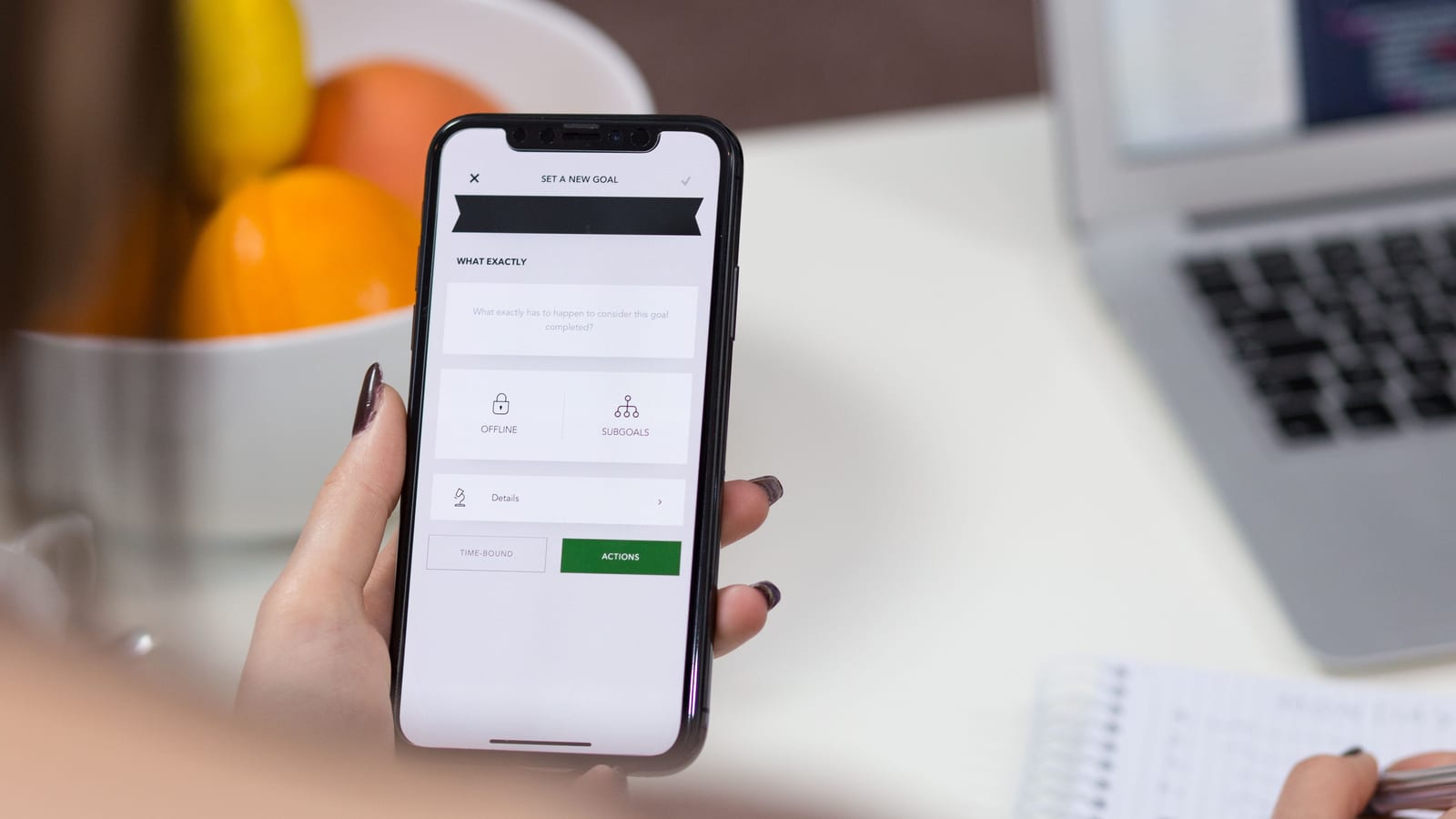 Close-up of a hand holding a smartphone showing a simple app form titled Set a New Goal, with Offline and Subgoals options and a green Actions button, a laptop and notebook blurred on the desk behind