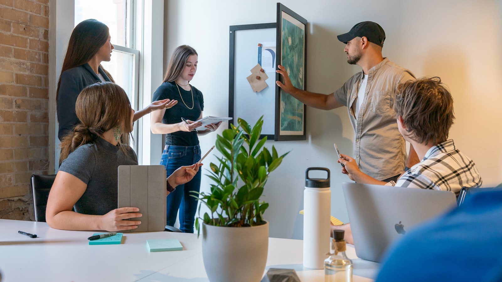 A small team in a bright brick-walled startup office, one member levelling a framed picture on the wall while colleagues discuss notes and a tablet around a white meeting table