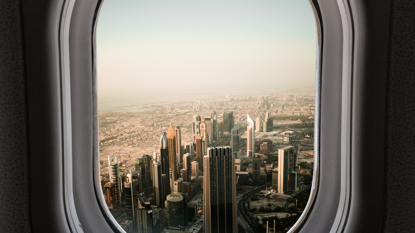 The Dubai skyline along Sheikh Zayed Road seen from inside an aircraft cabin, the city's towers and coastline framed by the rounded grey plastic surround of a passenger window in hazy golden light