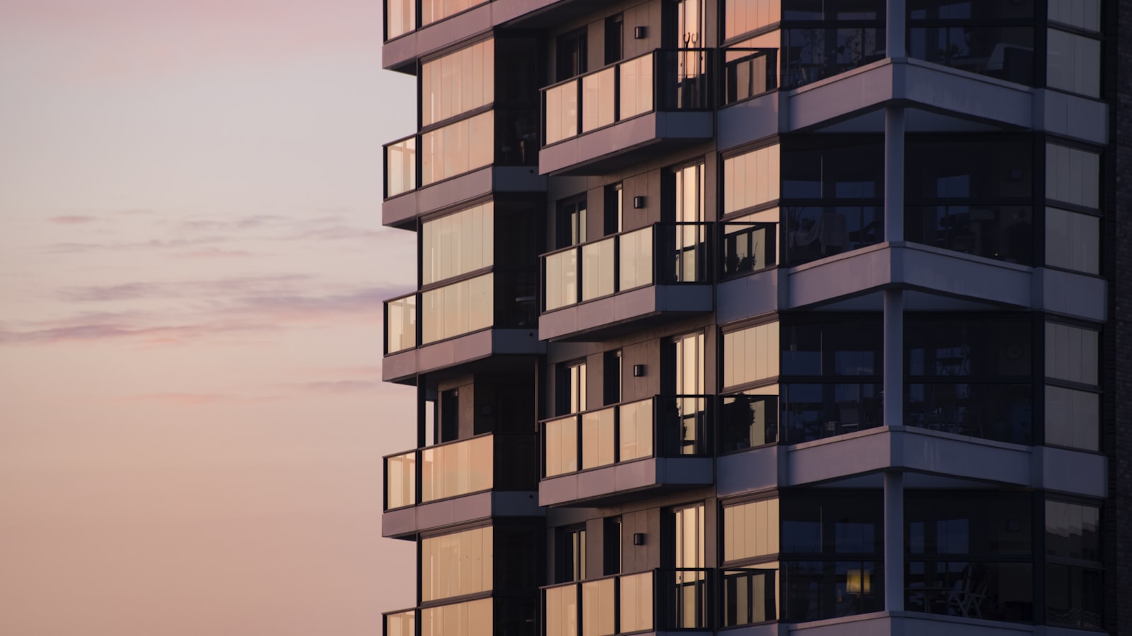 A modern residential apartment tower at dusk, its glass-fronted balconies and warmly lit apartment windows stacked floor by floor against a soft pink sky