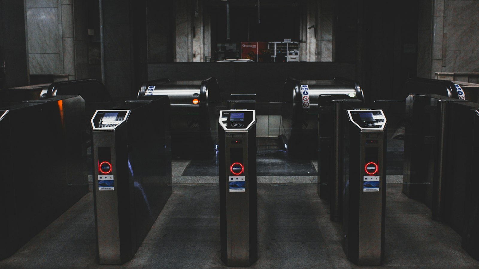 A row of black access-control speed gates with contactless card readers and glowing red no-entry indicators, standing in a dimly lit marble-clad building lobby