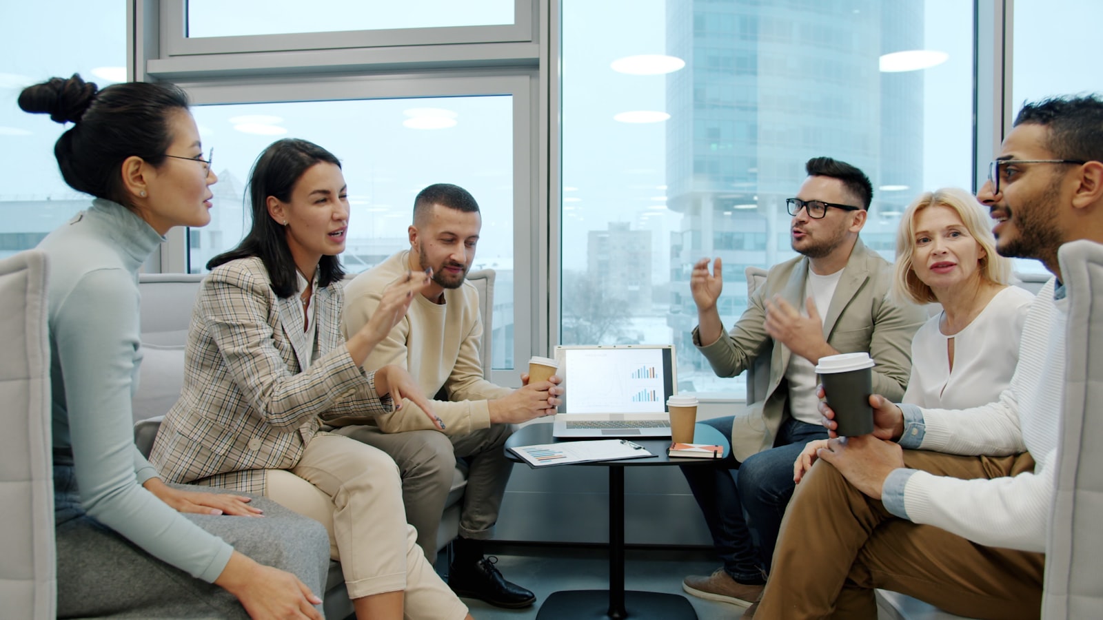 Six professionals of different nationalities sitting together in a bright modern lounge with high-rise windows behind them, talking over coffee cups and a laptop showing charts