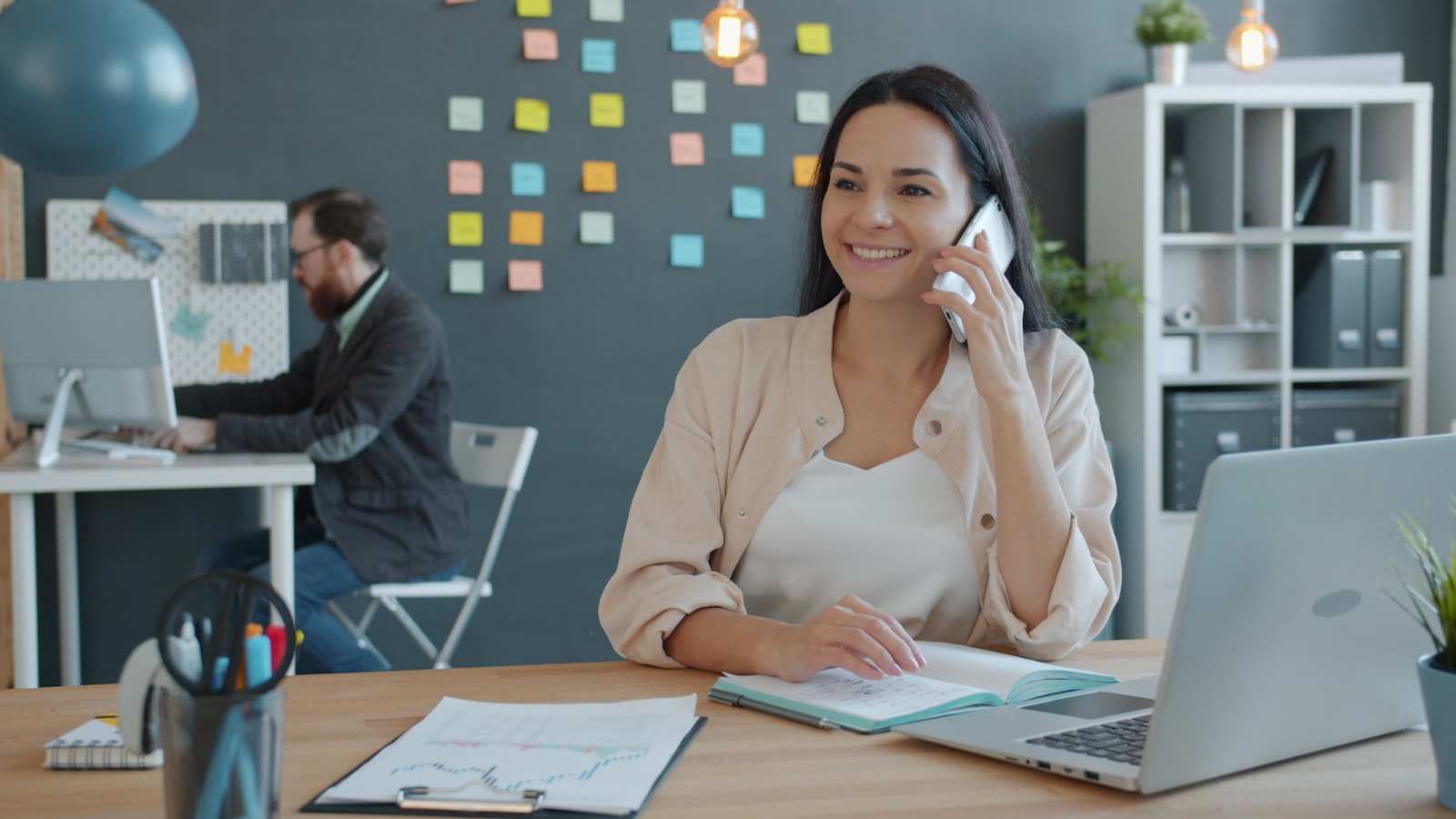A smiling real estate agent taking a client call at her desk with an open notebook, laptop, and performance charts while a colleague works in the background
