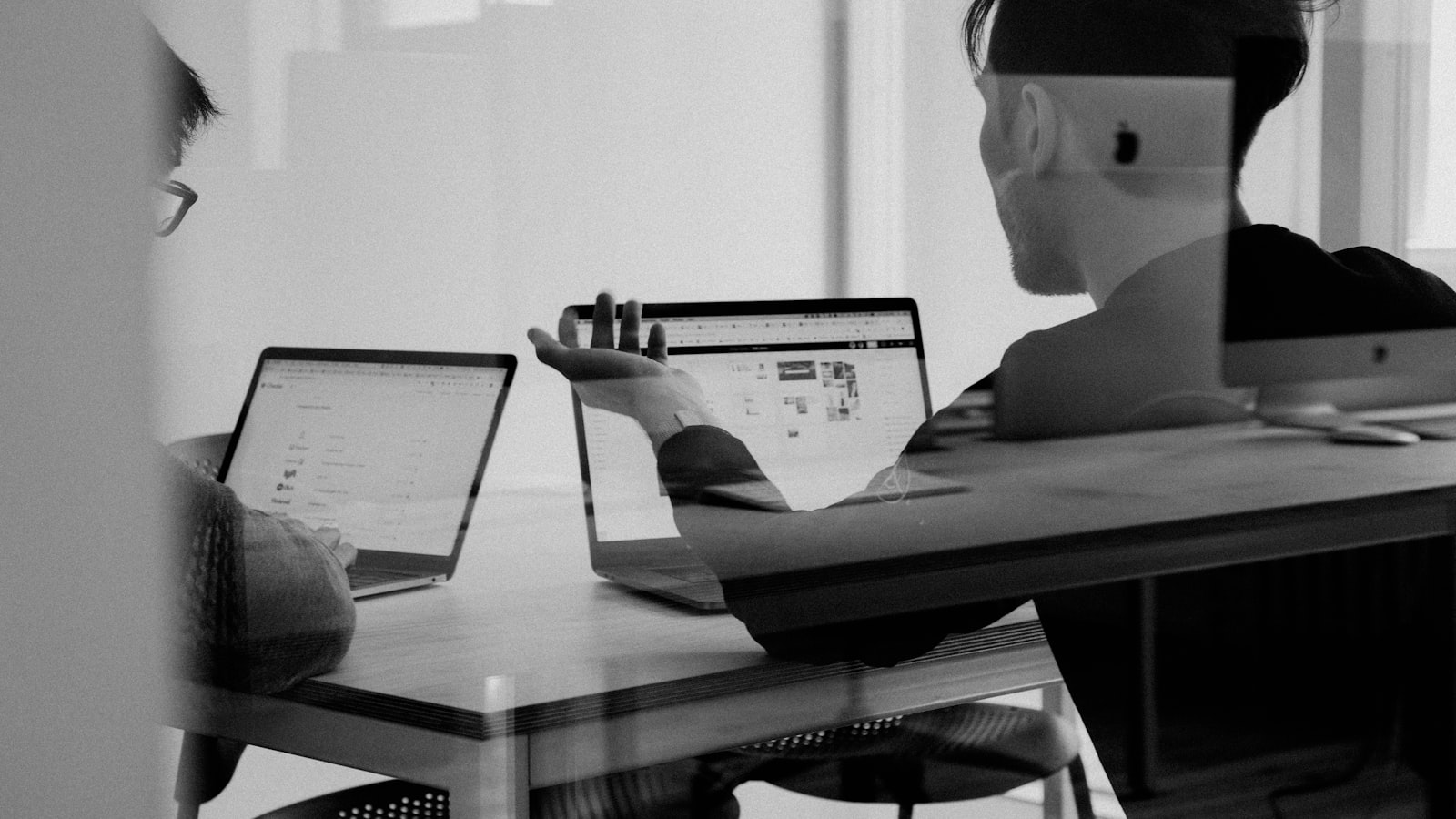 Black-and-white photo of two colleagues at a desk weighing software options across two open laptops and a desktop monitor, one gesturing mid-discussion
