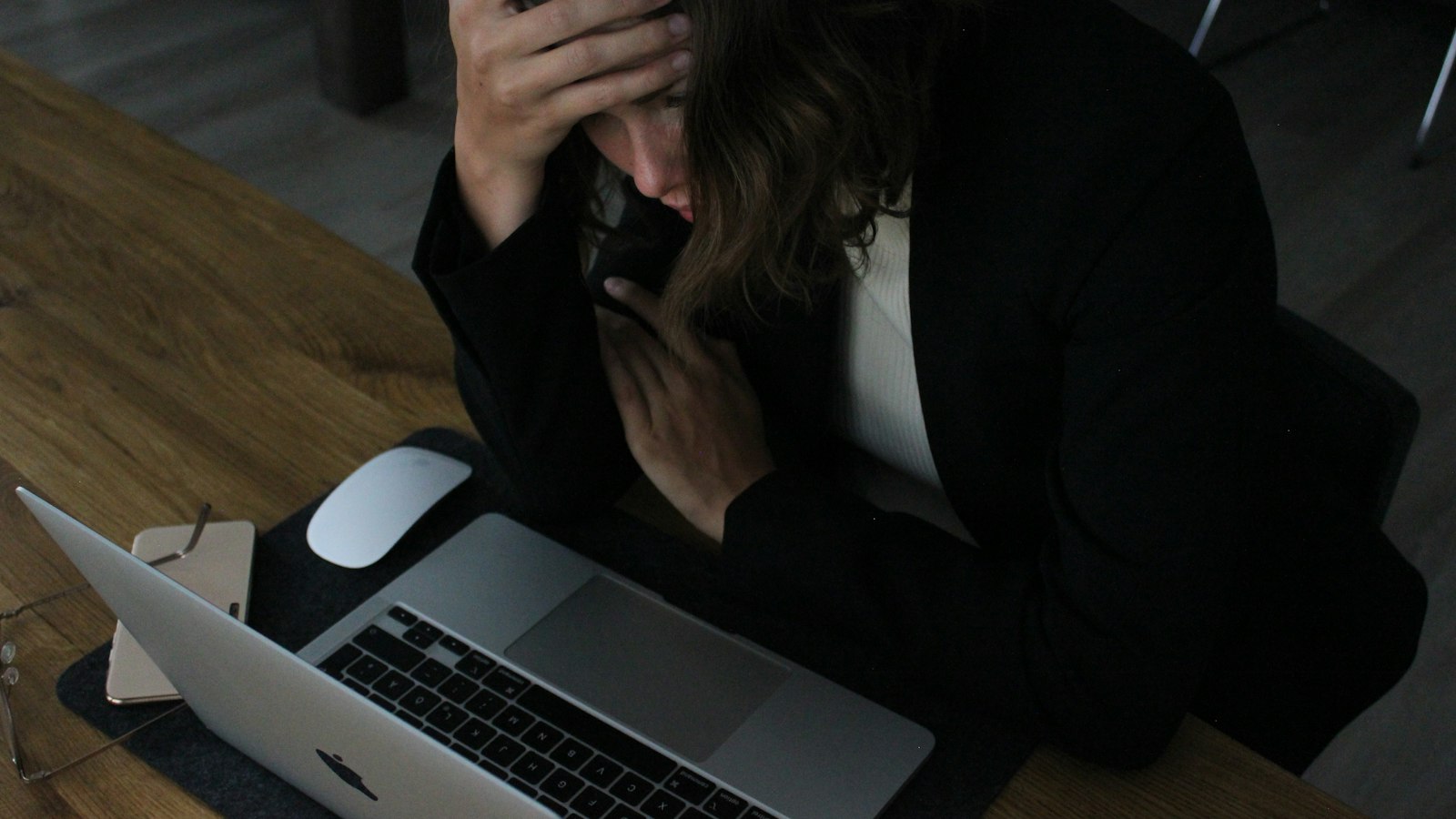 Overhead view of a woman in a dark blazer slumped over an open laptop in dim light, one hand gripping her forehead in visible frustration beside a mouse and folded glasses
