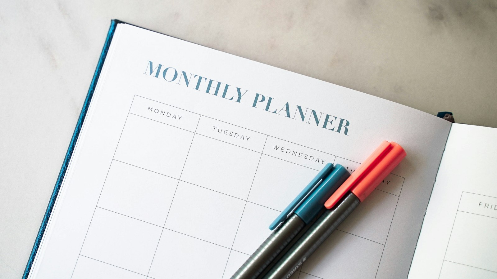 An open hardback monthly planner on a marble desk, its blank Monday to Friday grid ruled across the page, with two capped fineliner pens resting on it