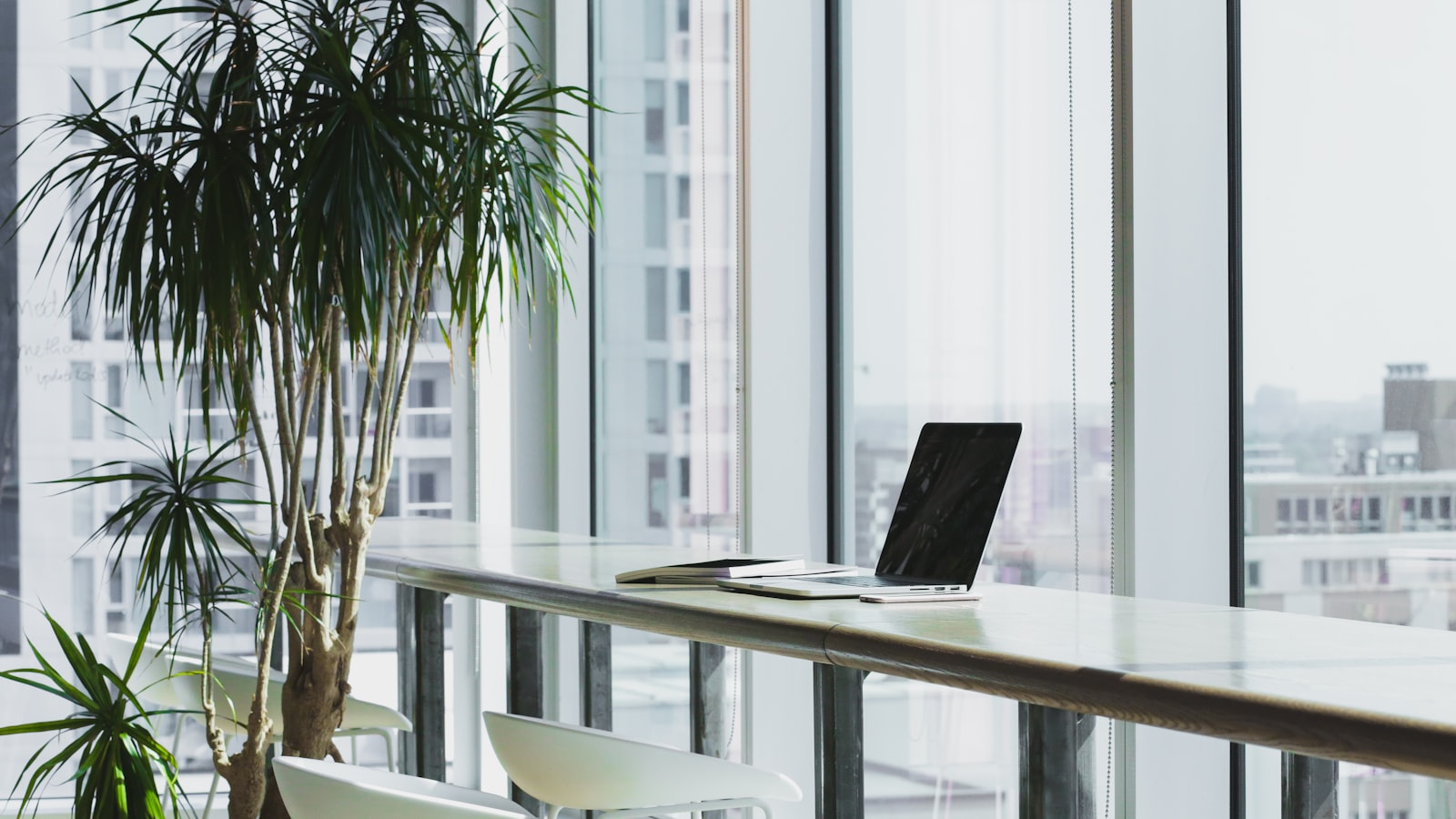 A laptop open on a wooden counter beside floor-to-ceiling windows overlooking a modern city skyline in a bright office