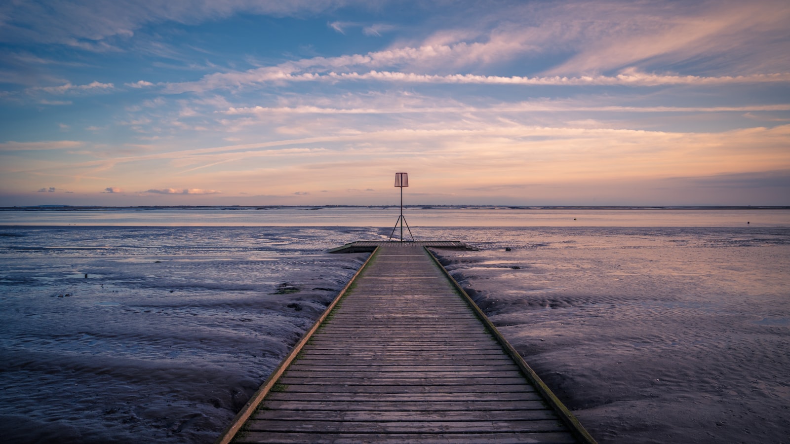 An empty timber walkway running straight away from the viewer across wide tidal flats to a lone metal marker post standing at its far end, under a pink and blue dusk sky