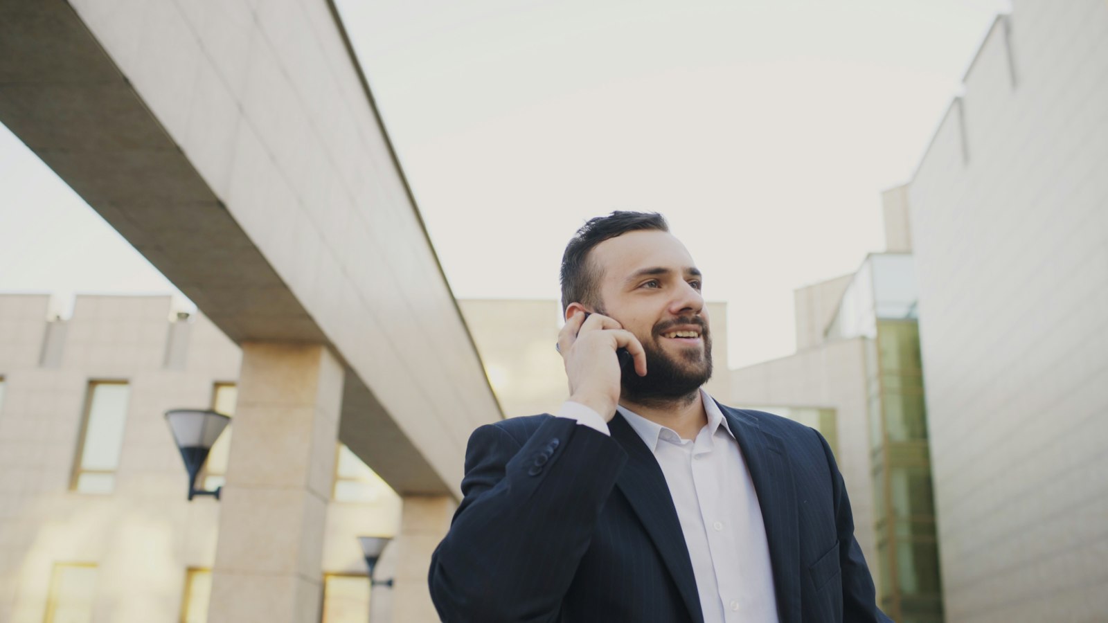 A bearded man in a dark suit and open white shirt taking a call on his mobile phone outdoors, shot from a low angle against the stone and glass facades of a modern building complex