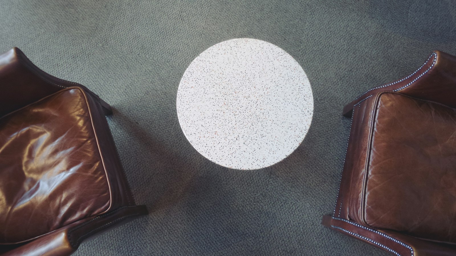Overhead view of two brown leather armchairs facing each other across a small round speckled-stone table on grey carpet, an empty two-seat negotiation setting