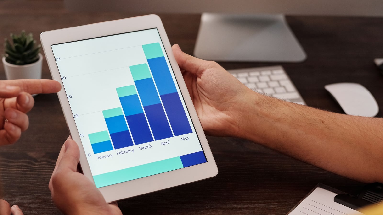 Close-up of two hands holding a white tablet that shows a stacked blue and teal bar chart climbing month by month from January to May, while a second person's finger points at the screen over a dark wooden desk with a small succulent, a keyboard, a mouse and a desktop monitor stand blurred behind