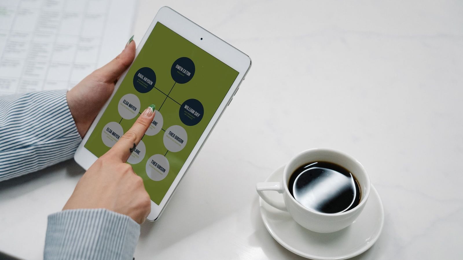 Overhead view of a hand pointing at a white tablet whose green screen shows a network of connected circular contact nodes with placeholder names, held over a white marble desk beside a cup of black coffee on a saucer