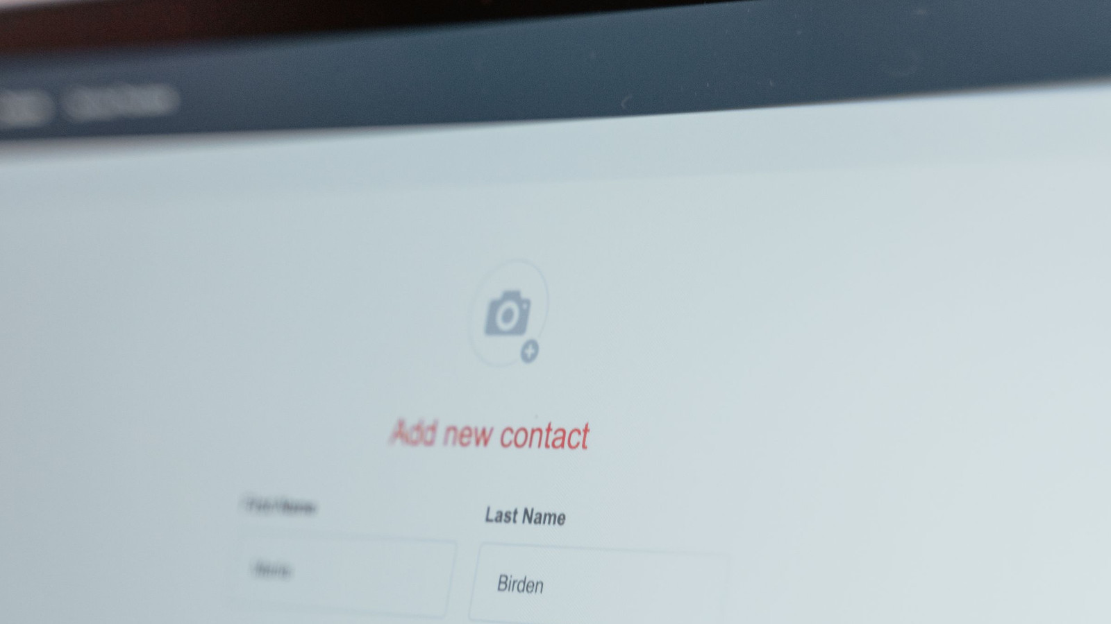 Close-up of a laptop screen showing a CRM's Add new contact form, with a camera icon for the contact photo above red Add new contact text and First Name and Last Name fields, the top bezel and menu bar softly blurred
