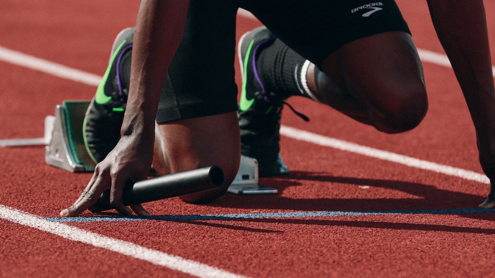 A close-up of a sprinter crouched in starting blocks on a terracotta running track, fingertips pressed to the white start line beside a black relay baton lying on the ground