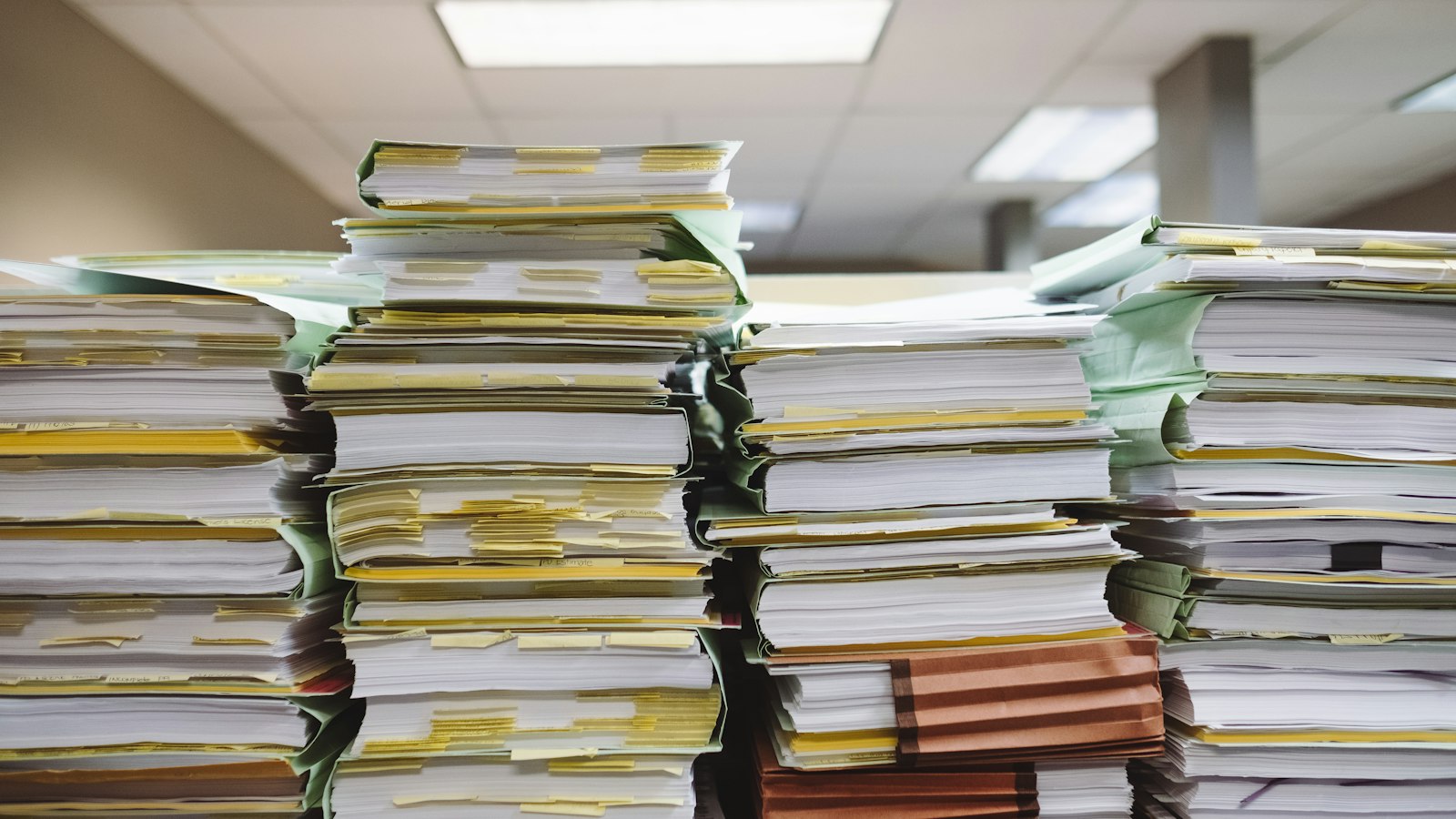 Towering stacks of paper files and tabbed documents piled high on a desk in an office, lit by overhead fluorescent panels