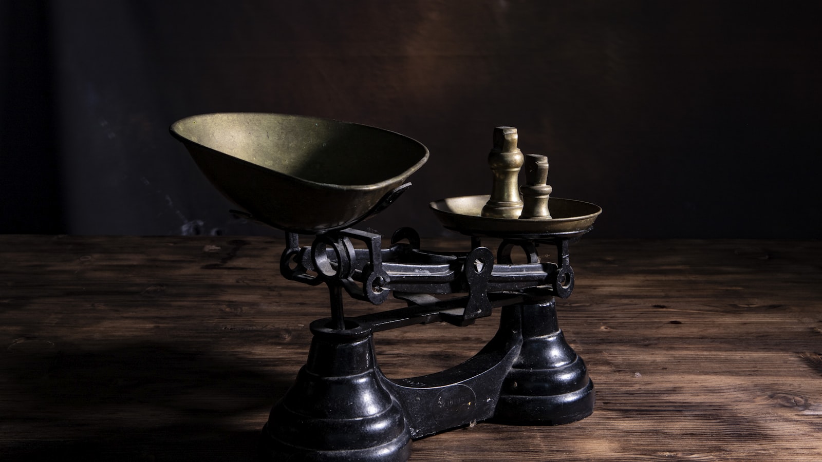 A black cast iron balance scale with brass weights stacked on its smaller pan, standing on a dark weathered wooden table against a deep shadowed background